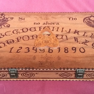 May include: A wooden Ouija board with a planchette in the centre. The board has the letters of the alphabet, numbers 1-9 and 0, and the words "Si", "No", "Hola", and "Adios".