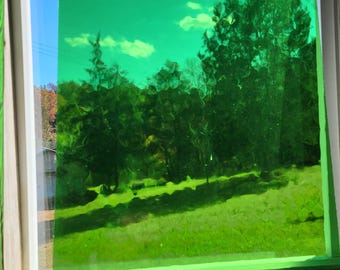 May include: A view through a window with a green tint. The scene outside shows a green grassy area, trees, and a blue sky with clouds. The window frame is white.
