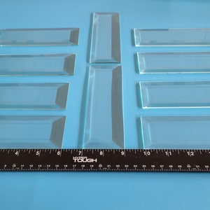 May include: Nine clear glass tiles with beveled edges, arranged in a symmetrical pattern on a blue surface. A ruler is placed at the bottom of the image for scale.
