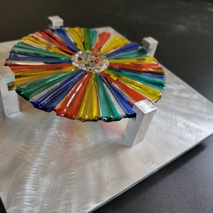 May include: A colorful glass sunburst design with a central mosaic, mounted on a silver metal base with four supports.