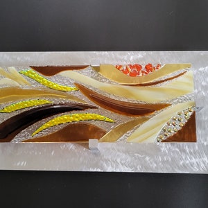May include: Abstract wall art featuring a mosaic of glass pieces in shades of brown, yellow, and orange. The glass pieces are arranged in a flowing, organic pattern, creating a sense of movement and energy. The artwork is framed in a silver metal frame.