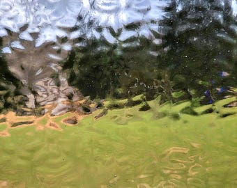 May include: A textured glass pane with a distorted view of a green lawn, trees, and a cloudy sky. The glass has a rippled surface, creating a blurred and abstract effect. The colors are muted, with greens, browns, and blues dominating the scene.