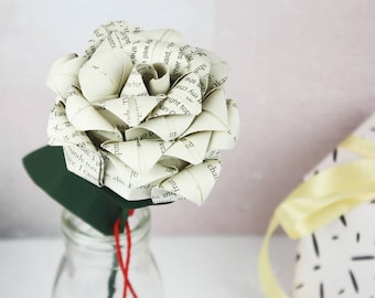 May include: A white paper rose made from the pages of a book, with a green stem and red twine, in a clear glass vase. The rose is sitting on a white surface.