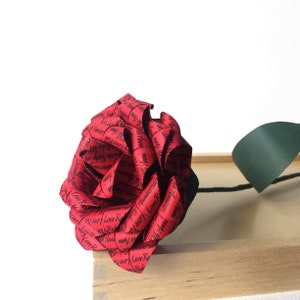 May include: A handcrafted red paper rose with black text, including the words "Love Ruby 40 Anniversary," is displayed. The rose is set against a light wooden box and a white background, with a green paper leaf.