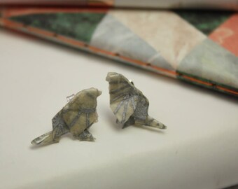 May include: A pair of origami bird earrings made from patterned paper. The birds are silver and gray with a white background. The earrings are a delicate and unique piece of jewelry.