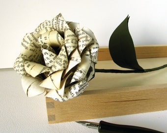 Könnte beinhalten: Eine Papierrose, die aus den Seiten eines alten Buches gefertigt wurde, mit einem grünen Stiel und Blättern. Die Rose liegt auf einer Holzoberfläche mit einem schwarzen Stift daneben.