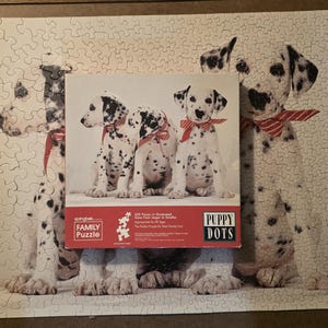 May include: A completed jigsaw puzzle featuring a Dalmatian dog design with a red bow tie. The puzzle box shows three Dalmatian puppies with red bow ties. The puzzle is titled "Puppy Dots" and is a "Family Puzzle".