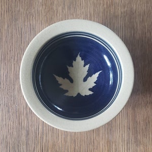 May include: A small ceramic bowl with a blue glaze and a white maple leaf design. The bowl has a beige rim.