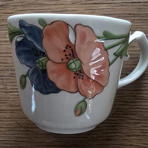 May include: White ceramic teacup with a handle, decorated with a large orange and blue floral design. The flowers have green stems and leaves. The cup is on a wooden surface.