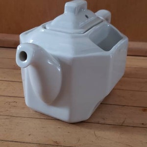May include: White ceramic teapot with a square body and a lid with a small spout. The teapot has a handle on the side and a small opening on the top for pouring.