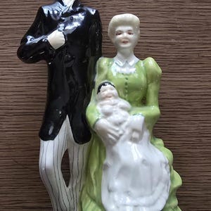 May include: A ceramic figurine depicting a family of three. The father figure is in a black suit with pinstriped pants. The mother figure wears a green dress and holds a baby wrapped in white. The figurines are on a brown surface.