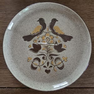 May include: A round, speckled ceramic serving platter with a brown and yellow bird and floral design. The platter has a silver handle and a brown rim. The design features two birds facing each other above a flower.