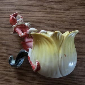May include: A vintage ceramic figurine featuring a whimsical elf figure leaning over a tulip-shaped vase. The elf wears a red outfit with a white collar and black shoes. The vase is yellow with red accents.