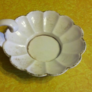May include: White ceramic saucer with a scalloped edge and a single handle. The saucer has a slightly aged look with a subtle brown tint.