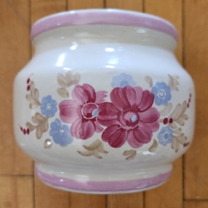 May include: A white ceramic jar with a pink rim and floral design. The jar features pink and blue flowers with brown leaves.