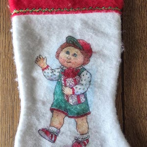 May include: White Christmas stocking with a red cuff and a green and gold trim. The stocking features a cartoon illustration of a child holding a wrapped gift. The child is wearing a green outfit and a red hat.