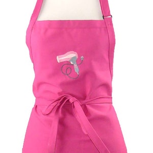 May include: A bright pink apron with a hairdryer design embroidered on the front. The apron has two pockets, a waist tie, and an adjustable neck strap. This apron is designed for use in a salon or at home.