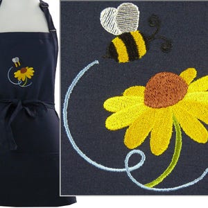May include: Navy blue apron with an embroidered design of a yellow sunflower and a bee. The bee has black and yellow stripes and white wings. The apron has a tie at the waist and an adjustable neck strap.
