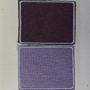 May include: Two rectangular fabric patches with rounded corners and stitched borders. The top patch is a deep dark purple with a smooth texture, while the bottom patch is a lighter lavender purple with a visible ribbed or waffle-knit texture.