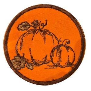 May include: A circular embroidered patch featuring a bright orange background with a dark brown stitched border. The design depicts two pumpkins in dark brown thread, with one larger pumpkin on the left and a smaller one on the right.