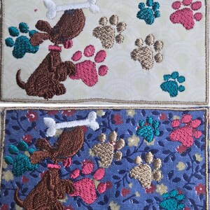 May include: Embroidered patches featuring a brown dog holding a white bone, surrounded by paw prints in teal, pink, and tan. The patches have a cream background with a subtle pattern, and a blue floral background.