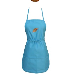 May include: A light blue apron featuring a central embroidered graphic of an artist's palette with colorful paint dabs and brushes. The apron includes a neck strap, a waist tie, and two large front pockets at the bottom for storage and utility.