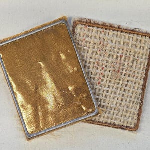 May include: Two rectangular patches with rounded corners. One patch features a smooth, metallic gold surface with a silver-colored stitched border. The second patch displays a coarse, beige woven fabric texture with a brown stitched border.