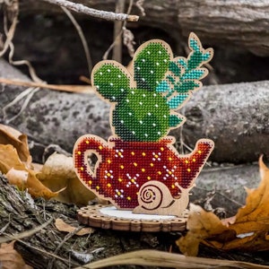 Può includere: Un ritaglio di legno di una teiera rossa con un cactus verde e una foglia verde che cresce da essa. Una lumaca è sul lato della teiera. La teiera è seduta su una base di legno.