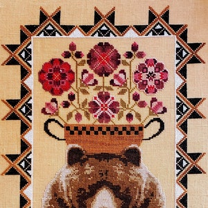 May include: Cross-stitch artwork featuring a bear with a floral arrangement. The bear is brown and tan, with a floral arrangement of red and pink flowers in a brown pot. The artwork is framed with a geometric border in black, white, and brown.