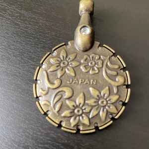 May include: A gold-toned metal cabinet knob with a floral design and the word "JAPAN" inscribed on the front. The knob has a small, round handle on the top.