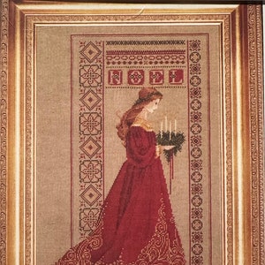 May include: A framed cross-stitch embroidery depicting a woman in a red dress holding a wreath with candles. The embroidery is surrounded by a gold frame with ornate details. The text "NOEL" is visible in the upper portion of the embroidery.
