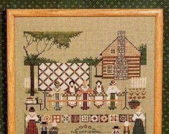 May include: Cross-stitch pattern featuring a log cabin with a gathering of people outside. The scene includes a quilt hanging on a fence, a table with food, and the text "The Gathering".