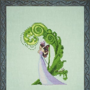 May include: Cross-stitch artwork featuring a woman in a lavender gown playing a harp beneath a large, green, fern-like structure. The piece is framed in a decorative, dark gray border, set against a light green background. The woman has brown hair and is wearing a crown.