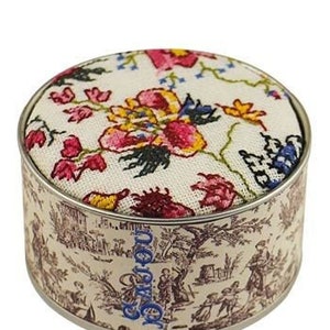 May include: Round pin cushion with a floral design on top. The cushion is set in a metal tin with a toile pattern. The floral design features pink, yellow, and blue flowers on a cream background.