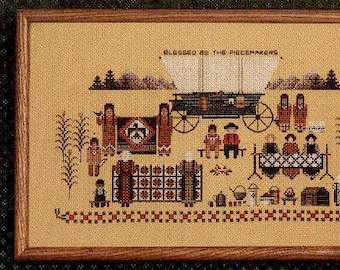 May include: A cross-stitch pattern featuring a covered wagon with the text "Blessed be the peacemakers" above it. The scene depicts a group of people in traditional clothing gathered around a table with a quilt in the background.