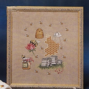 May include: Cross-stitch artwork in a gold frame with a bee theme. The design includes a beehive, honeycomb, bees, a jar of honey, and a flower. The background is a tan fabric.