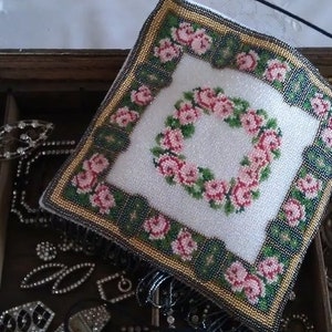 May include: A square beaded pincushion featuring a white center with a pink rose wreath, surrounded by a border of green and gold beads with pink rose accents. The pincushion is placed atop a wooden box filled with various metal jewelry pieces.