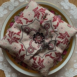 Puede incluir: Una almohada bordada en beige y rojo con un diseño floral, descansando sobre un plato con un patrón floral. Un par de tijeras antiguas se colocan encima de la almohada.