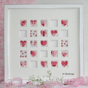 Puede incluir: Un marco blanco con un paspartú blanco y 16 diseños de punto de cruz en forma de corazón rojo y blanco. El marco está sobre una superficie rosa y blanca con flores blancas.