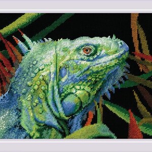May include: A framed cross-stitch of an iguana, showcasing a detailed close-up of its head and neck. The iguana is rendered in shades of green and blue, with a reddish-brown eye. The background features colorful foliage against a black backdrop.