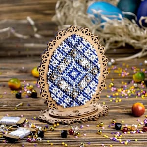 May include: Wooden Easter egg decoration with a blue and white beaded design. The egg is set in a wooden stand with a decorative edge. Colorful beads and Easter eggs are scattered around the base.