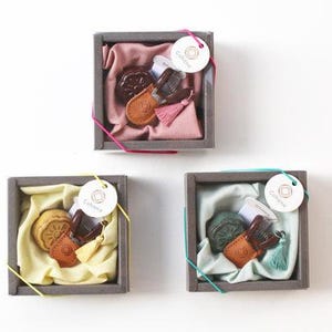 May include: Three square gift boxes with various contents. Each box contains a small leather item, a decorative item, and a tassel. The boxes are lined with fabric in pink, yellow, and teal. Each box has a tag attached.
