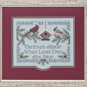 Puede incluir: Bordado de punto de cruz con fondo azul, que presenta dos cardenales rojos posados en ramas de pino con una casita para pájaros en el centro. El texto dice "Cardinals Appear When Loved Ones Are Near."