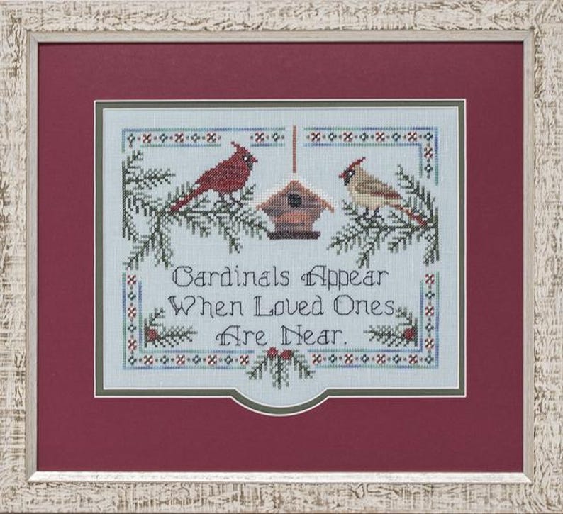 May include: A framed cross-stitch embroidery on light blue fabric featuring two cardinals, a birdhouse, and pine branches. The text reads, "Cardinals Appear When Loved Ones Are Near." The piece is set within a deep red mat and a textured white frame.