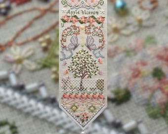 Puede incluir: Un detallado estandarte de punto de cruz con las palabras "Apple blossom" y los números "20" y "25". El estandarte presenta motivos florales y de mariposas, un árbol y un borde decorativo. Está rematado con un colgador de metal dorado y una borla.