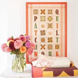 Könnte beinhalten: Ein gerahmtes Kreuzstichstück mit dem Wort "PATCHWORK", das in braunen Buchstaben ausgeschrieben ist. Jeder Buchstabe ist von einem anderen bunten Quiltblockmuster umgeben. Der Rahmen ist hellorange und der Hintergrund ist ein hellbeiger Stoff mit einem dezenten Karomuster. Eine weiße Steppdecke mit Blumenmuster liegt auf einem Tisch vor dem gerahmten Stück. Eine Vase mit rosa und orangefarbenen Dahlien steht auf dem Tisch links von der Steppdecke.