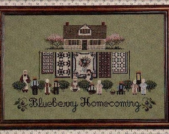 May include: Framed cross-stitch artwork featuring a house, quilts, and figures. The artwork is in a wooden frame. The text "Blueberry Homecoming" is stitched at the bottom. The color palette includes green, brown, pink, and white.
