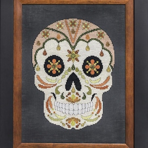 Puede incluir: Una calavera de azúcar blanca con diseños florales naranja, verde y amarillo. La calavera está enmarcada en un marco de madera negro y marrón.