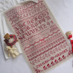 May include: A cream-coloured cross-stitch sampler with red embroidery. It displays different fonts, alphabets, and floral border designs. Two small teddy bears and a doll are positioned beside the sampler.