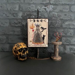 May include: A decorative display featuring a cross-stitch artwork of a queen with a black cat, a gold skull, and a wooden spool with scissors. The artwork is mounted on a stand. The background is a dark gray brick wall.